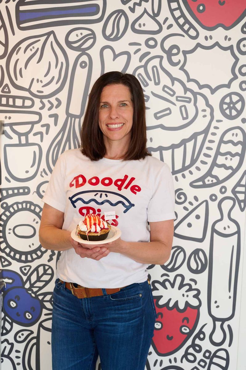 Anne Mullenax holding a Doodle Pie in front of the shop's doodle wall